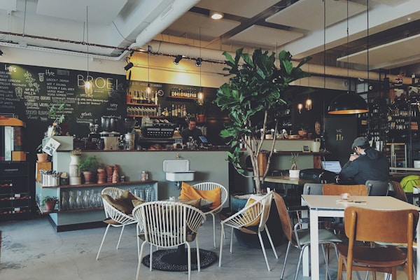 Cozy coffee shop interior with warm lighting, wooden tables, exposed brick walls, hanging plants, vintage coffee equipment, comfortable seating area, ambient atmosphere, professional photography